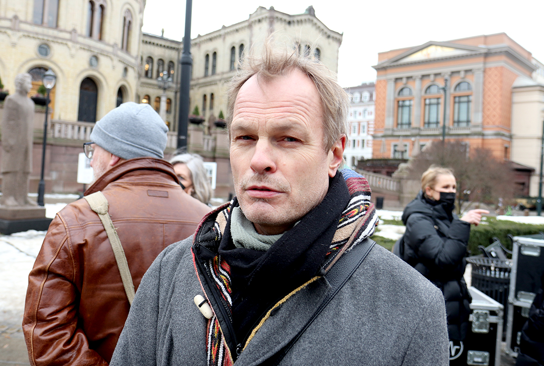 Mann med bustete blondt hår og grå jakke ser alvorlig ut foran Stortinget