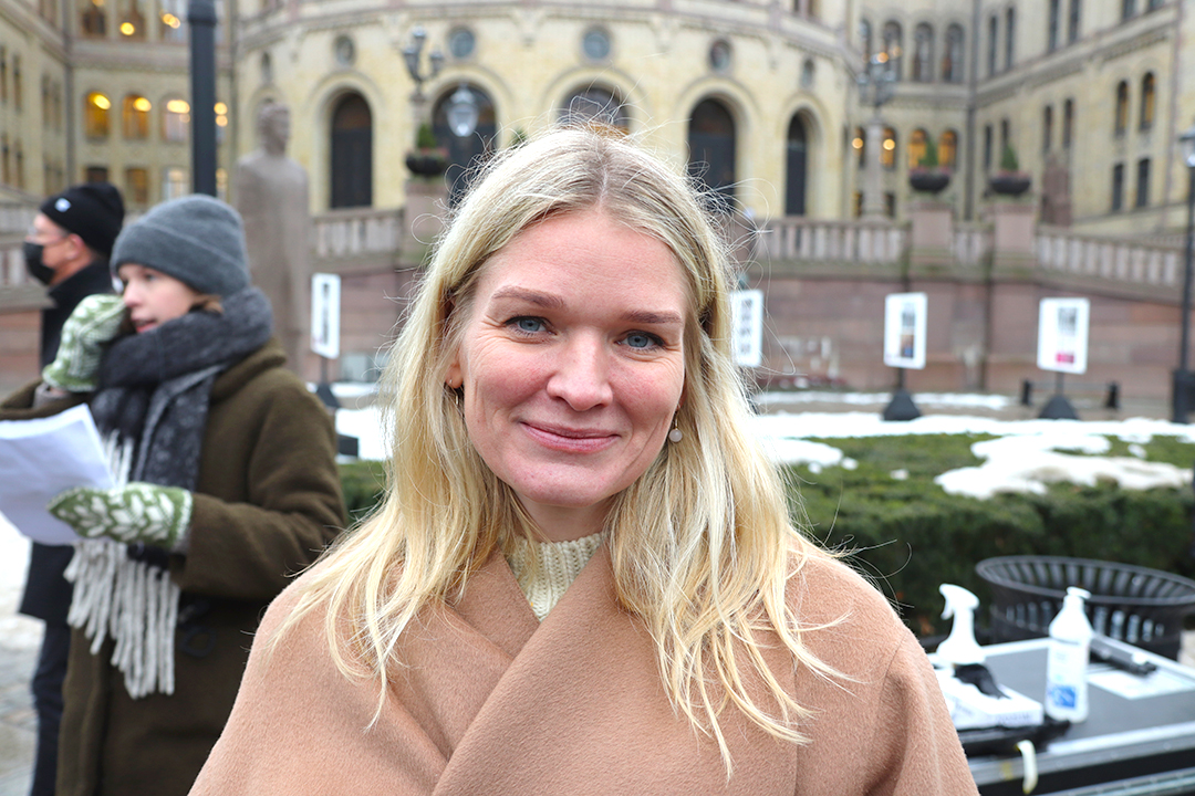 Kvinne med langt blondt hås og beige jakke smiler skjevt foran Stortinget