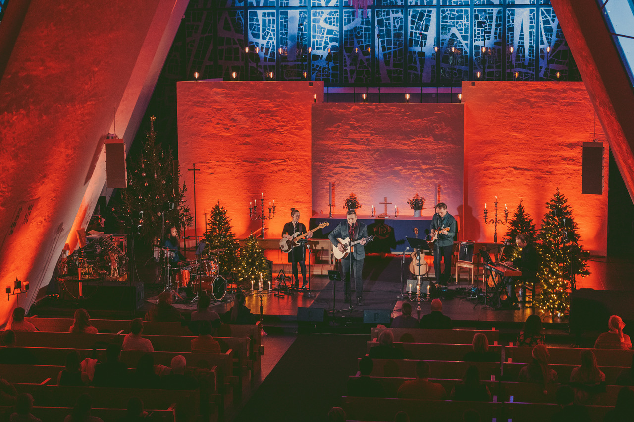 mann i dress med gitar og backingband spiller julekonsert i kirkerom med rød belysning