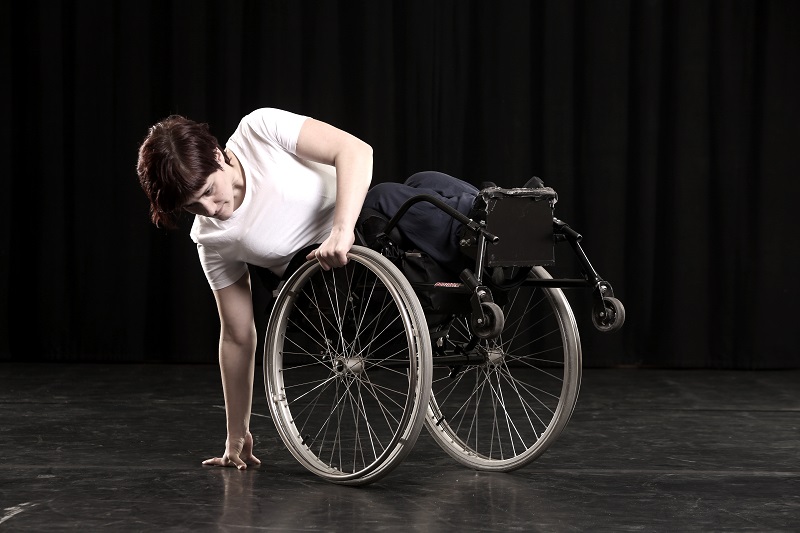 Kvinne i hvit t-skjorte og kort mørkt hår danser i rullestol