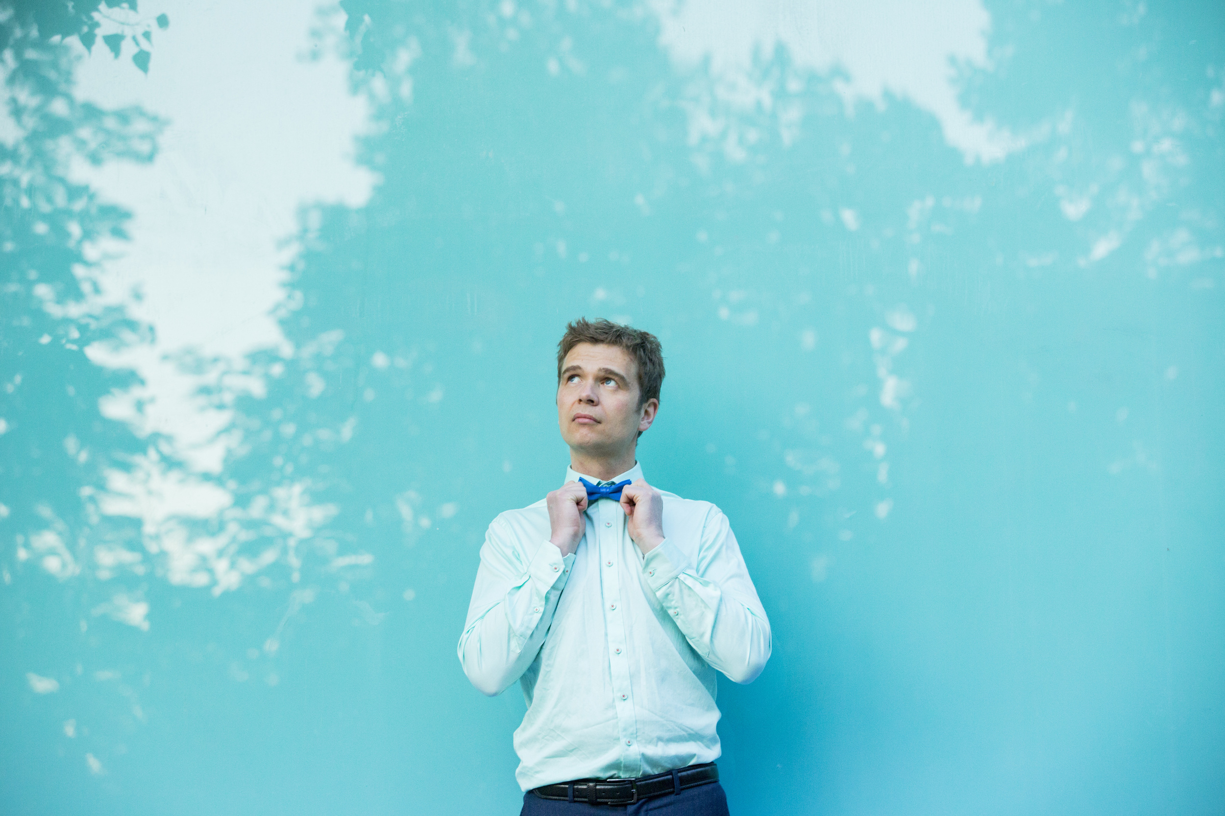 Mann i hvit skjorte, med blått slips lent mot turkis vegg