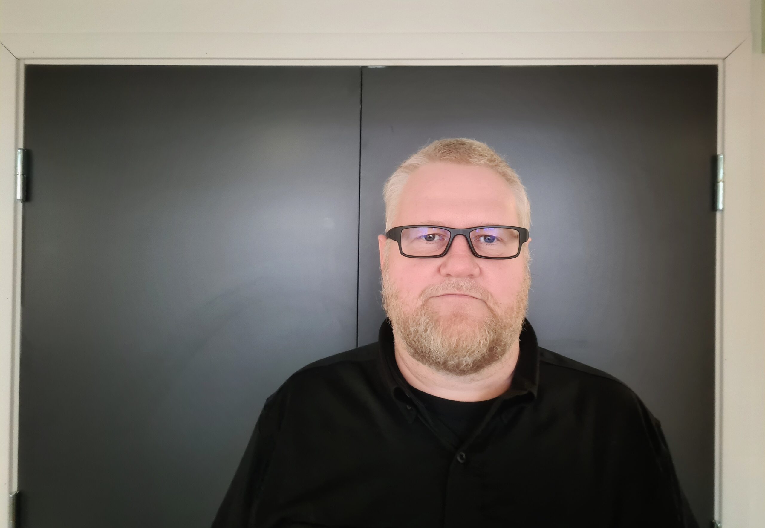 Mann med kort blondt hår, briller, skjegg og sort genser mot grå vegg
