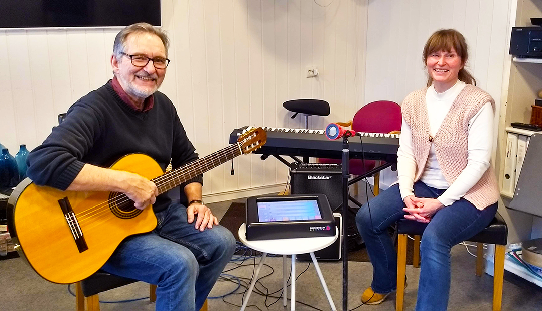 Mann smiler med gitar på fanget, kvinne på stol ved siden av smiler også