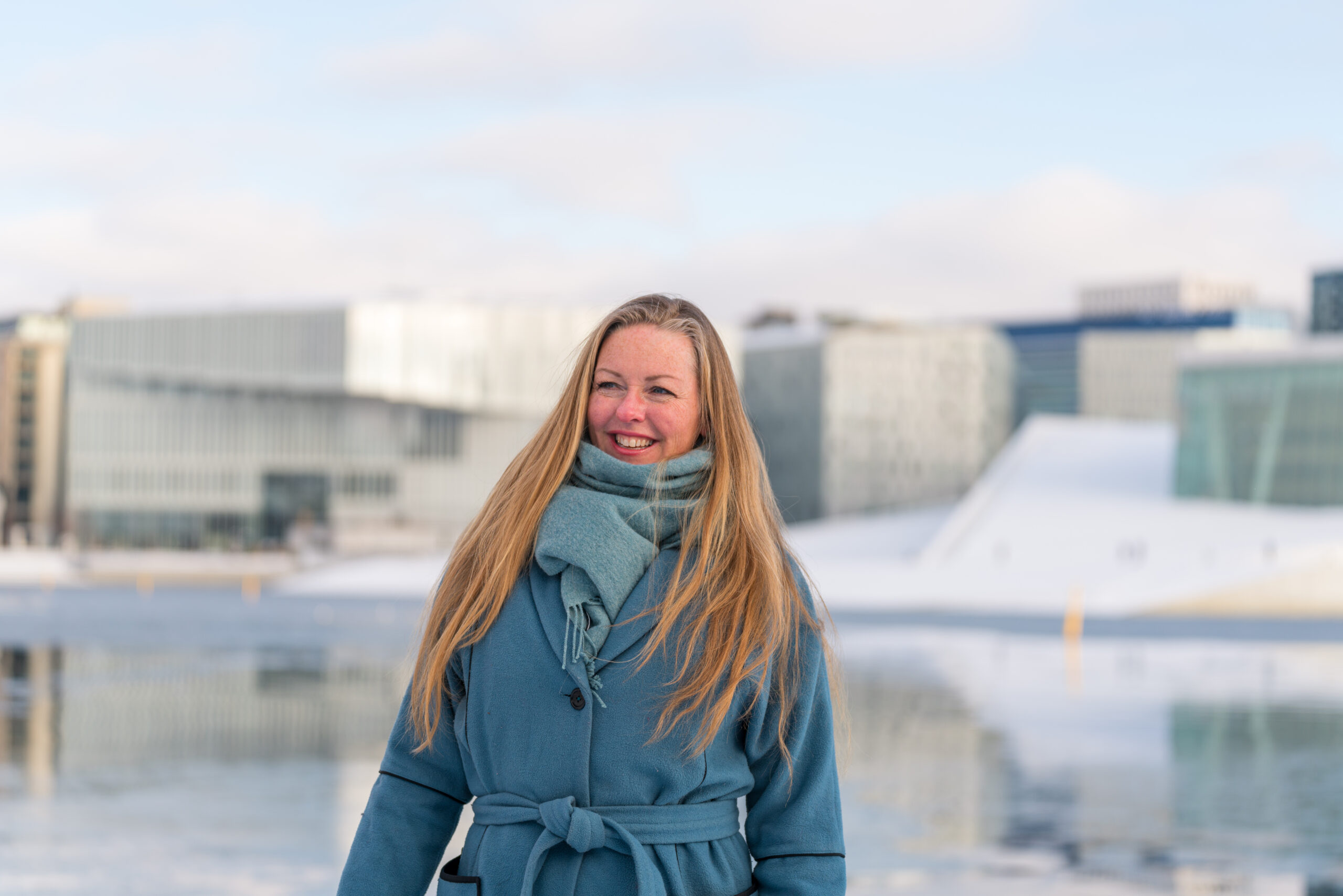 Kvinne med langt blondt hår og blå kåpe foran operahuset i Bjørvika